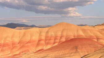 Rounded claystone mounds banded in gold, coral, and deep red rise in sunlit layered folds at the Painted Hills, with low blue-grey ridges on the horizon and a dark cloud band drifting across a pale blue sky in eastern Oregon.